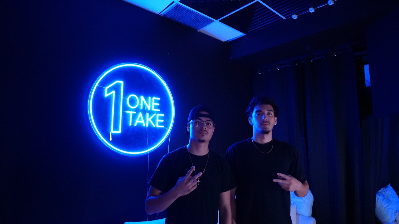 Two artists posing in front of the One Take neon sign